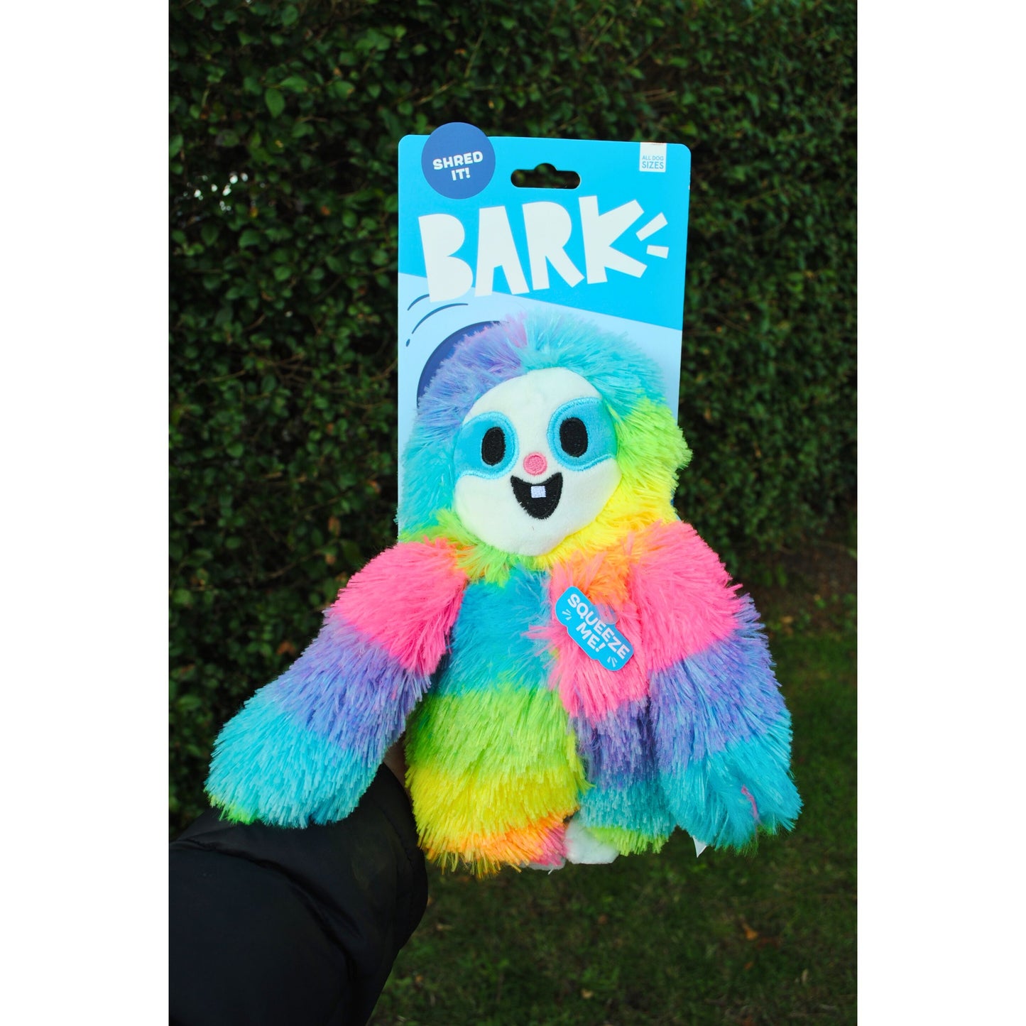 Colorful plush toy with 'Bark!' packaging held against a green hedge background