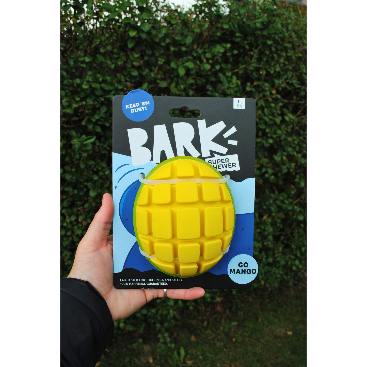 Yellow dog chew toy in packaging with 'Bark' branding against a green hedge background