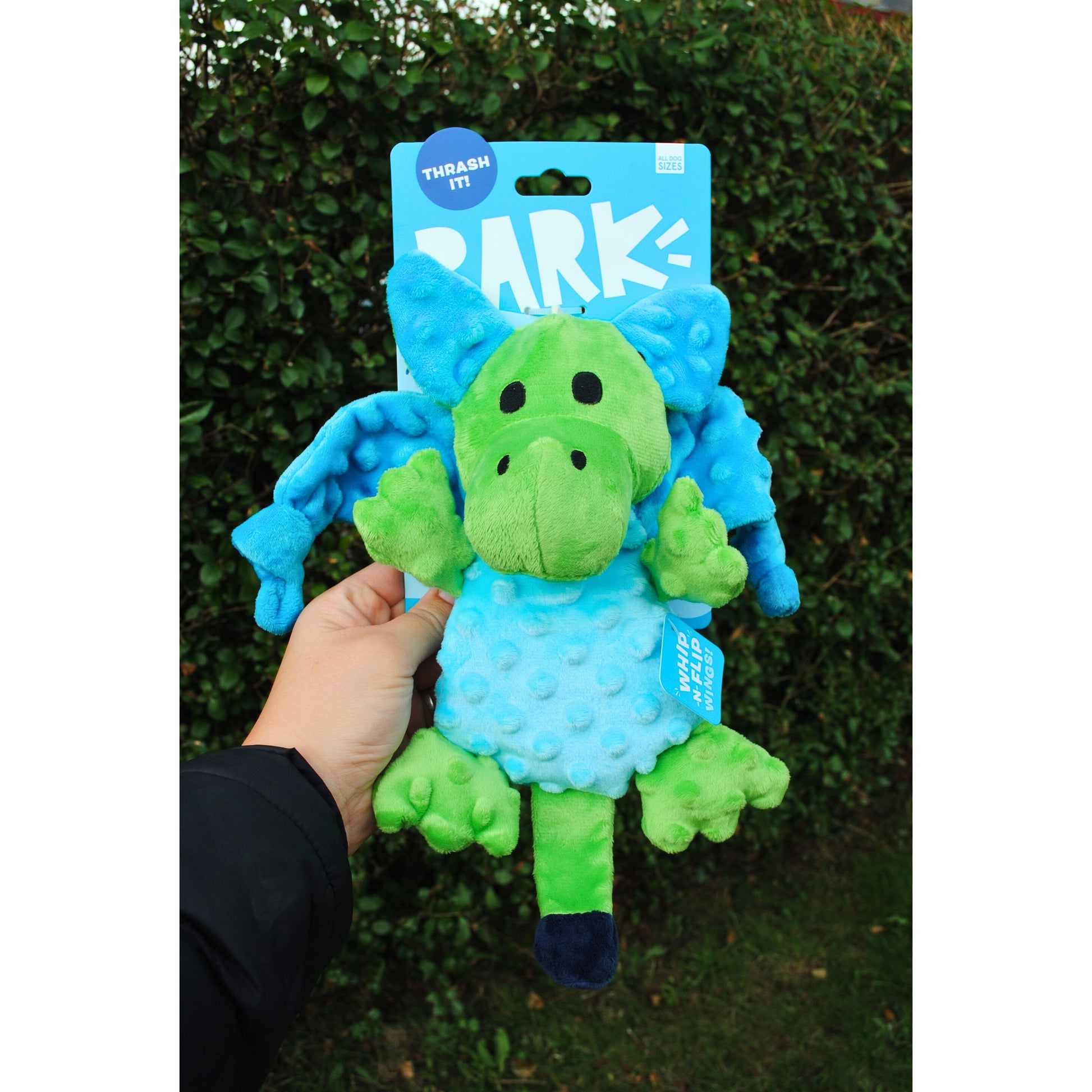 Green and blue plush toy with 'BARK' branding held by a hand against a green hedge background