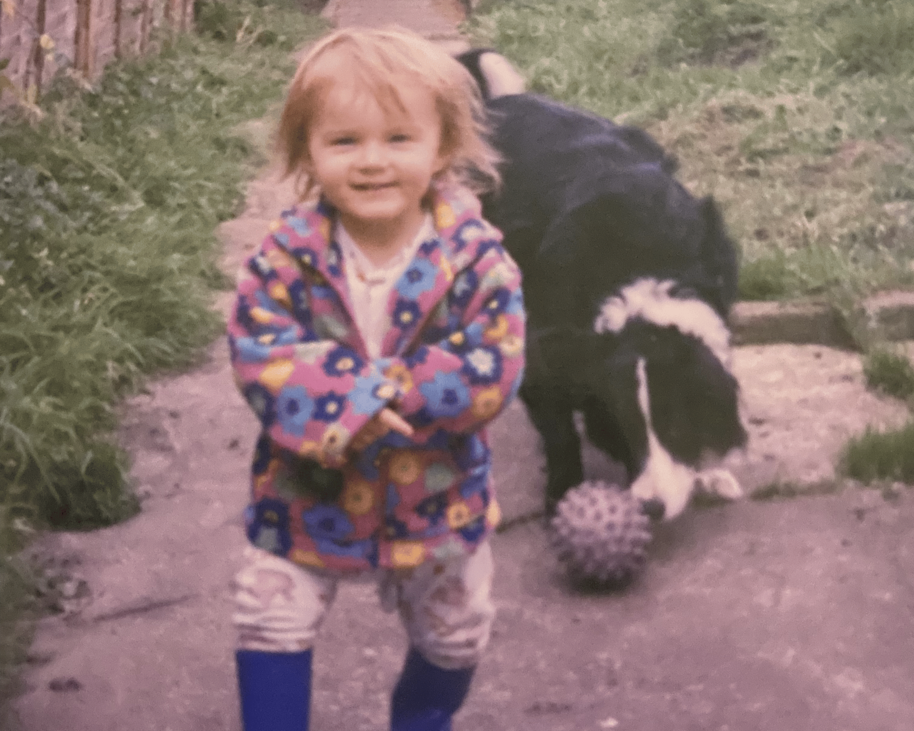 Child in a colorful coat and blue boots standing outdoors with a dog and ball in the background.